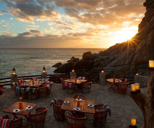 5 tables with placesettings and chairs on a stone patio with mountainside along the right side of the dining space and photo and the sea to the left. the sun is just setting behind the mountains, creating a sunburst effect over the stone