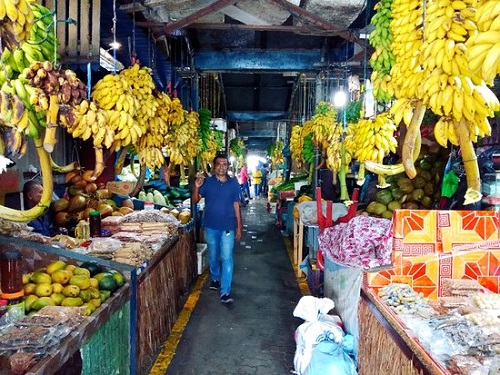 man in a blue shirt waving at camera between two long rows of market stalls stretching into the background of the photo. the stalls are full of local fruits, bananas and plantains hanging overhead
