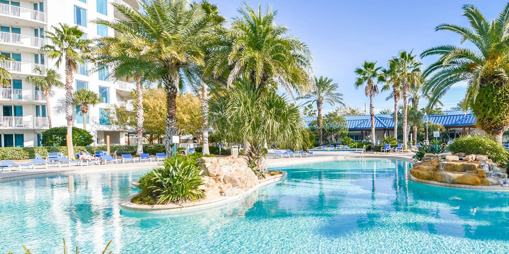 an irregularly shaped turquoise bottomed pool partially lined with palm trees with a false oasis within the pool